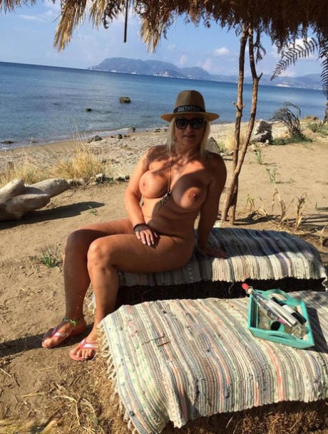 Thick blonde woman with big boobs wanders around a nude beach in a straw hat